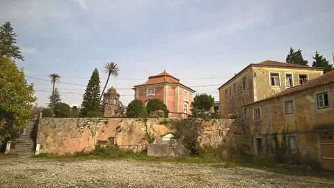 Edifícios históricos e muro de pedra na Quinta Real de Caxias, em Oeiras, com palmeiras e céu azul.