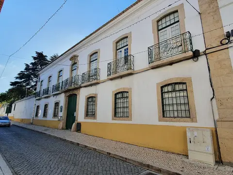Fachada branca e amarela de palácio com varandas de ferro forjado, junto a rua calcetada.