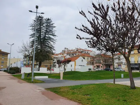 Jardim urbano com relvado, caminhos pavimentados e árvores, com casas da Azambuja ao fundo