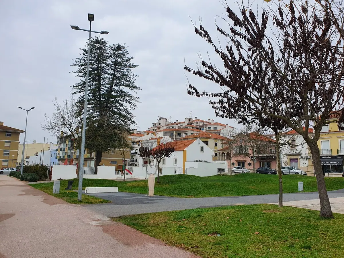 Jardim urbano com relvado, caminhos pavimentados e árvores, com casas da Azambuja ao fundo