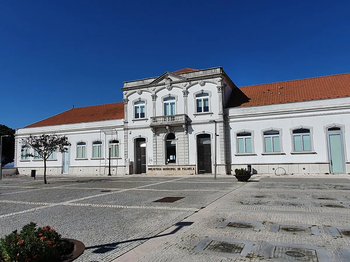 Fachada branca da Biblioteca Municipal de Palmela, com telhado de telha e largo calcetado em frente.