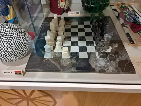 Tabuleiro de xadrez em pedra com peças esculpidas, em vitrina de museu junto a peças de Angola e Moçambique