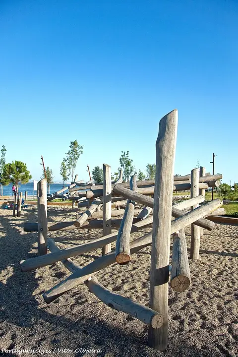 Estrutura de troncos de madeira num parque infantil de areia junto ao rio, sob céu azul