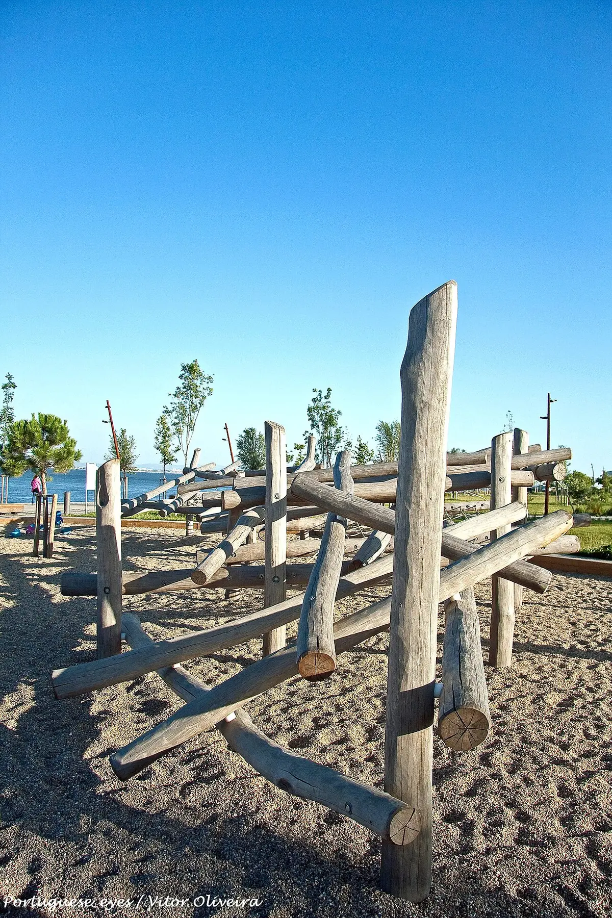 Estrutura de troncos de madeira num parque infantil de areia junto ao rio, sob céu azul