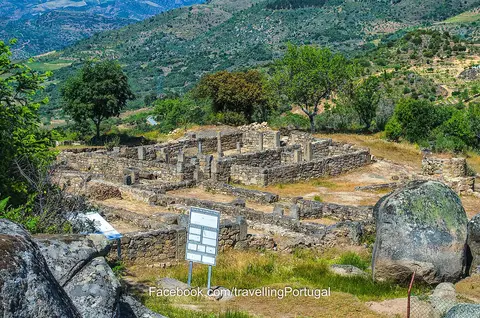 Ruínas arqueológicas de muros de pedra numa encosta verdejante, com painel informativo e vista para o vale