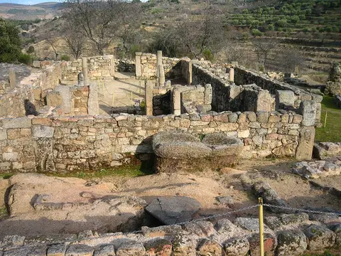 Ruínas arqueológicas em pedra de antiga igreja, com muros e vestígios, rodeadas por encostas verdes.