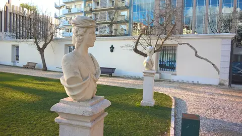 Bustos de pedra sobre pedestais num jardim relvado, com bancos e edifícios ao fundo