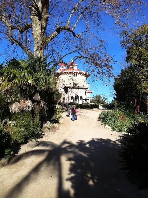 Caminho do Parque de Monserrate ladeado por vegetação, com o palácio ao fundo sob céu azul
