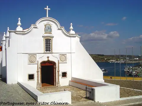 Fachada branca da Ermida de Nossa Senhora das Salvas, em Sines, com cruz e painel de azulejos, junto ao porto.