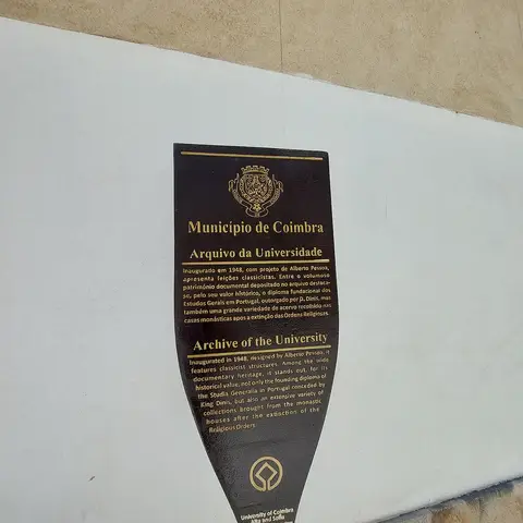 Placa informativa do Arquivo da Universidade de Coimbra, em metal escuro com letras douradas, numa parede branca