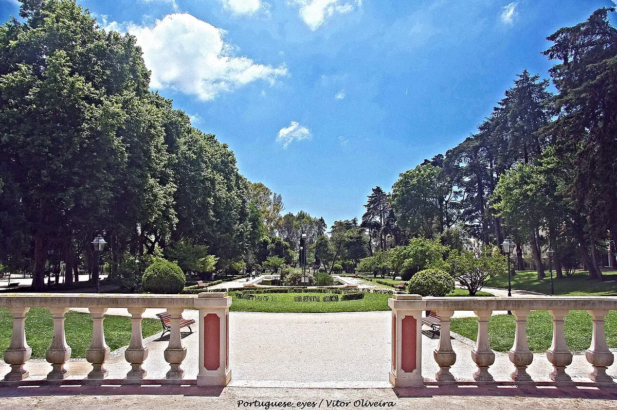 Jardim público com balaustrada de pedra, caminho central, canteiros, bancos e árvores altas.