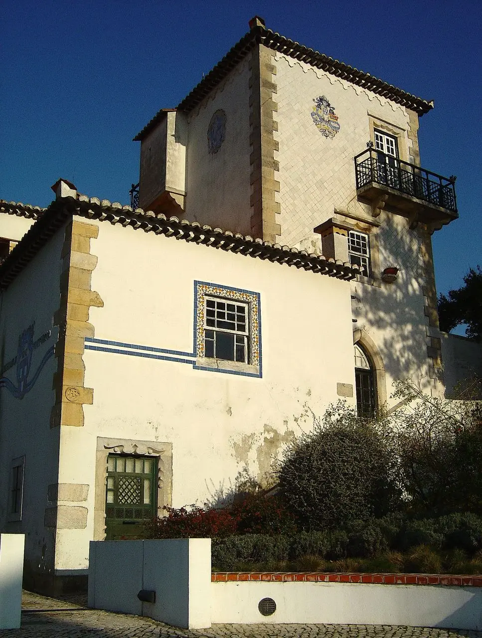 Fachada branca da Casa Roque Gameiro, com torre, azulejos e varanda de ferro, sob céu azul