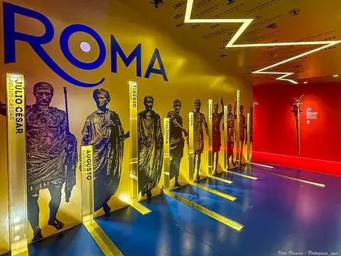 Sala expositiva do museu PO.RO.S com painéis de imperadores romanos e letreiro "Roma" em parede amarela