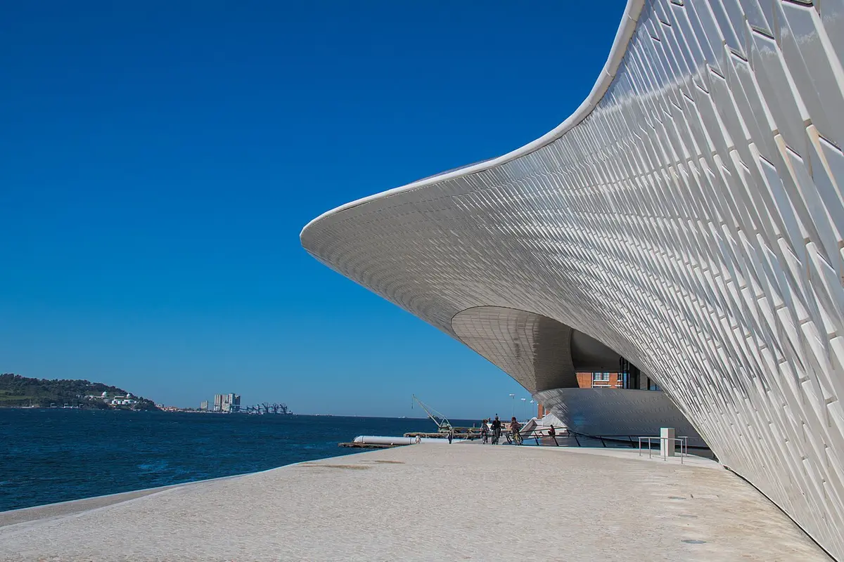 Edifício curvo e branco do MAAT junto ao rio Tejo, sob céu azul, com pessoas ao longe