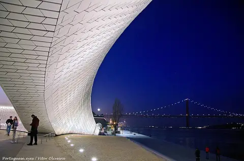 Edifício curvo e iluminado do MAAT à noite, junto ao Tejo, com a Ponte 25 de Abril ao fundo