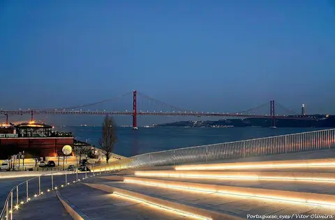 Cobertura em degraus iluminada do MAAT ao anoitecer, com o Tejo e a Ponte 25 de Abril ao fundo
