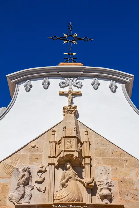 Frontão da Igreja Matriz de Moncarapacho com cruz de ferro e relevos em pedra sobre céu azul