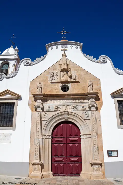 Fachada branca da Igreja Matriz de Moncarapacho com portal manuelino em pedra e portas vermelhas