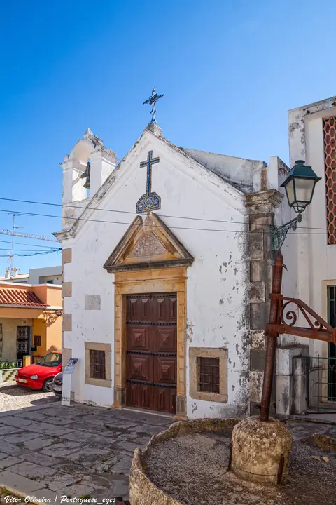 Fachada branca da Igreja do Santo Cristo em Moncarapacho, com portal de madeira e cruz de ferro.