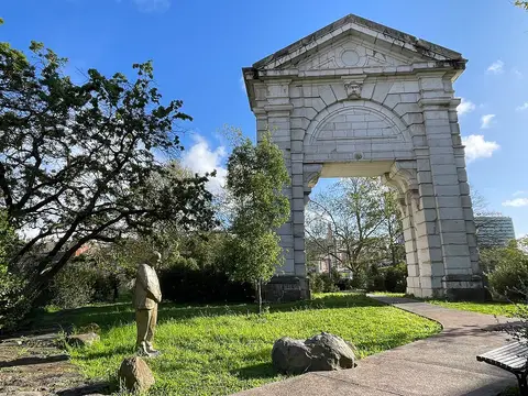 Arco de pedra monumental e estátua num jardim relvado com árvores, sob céu azul