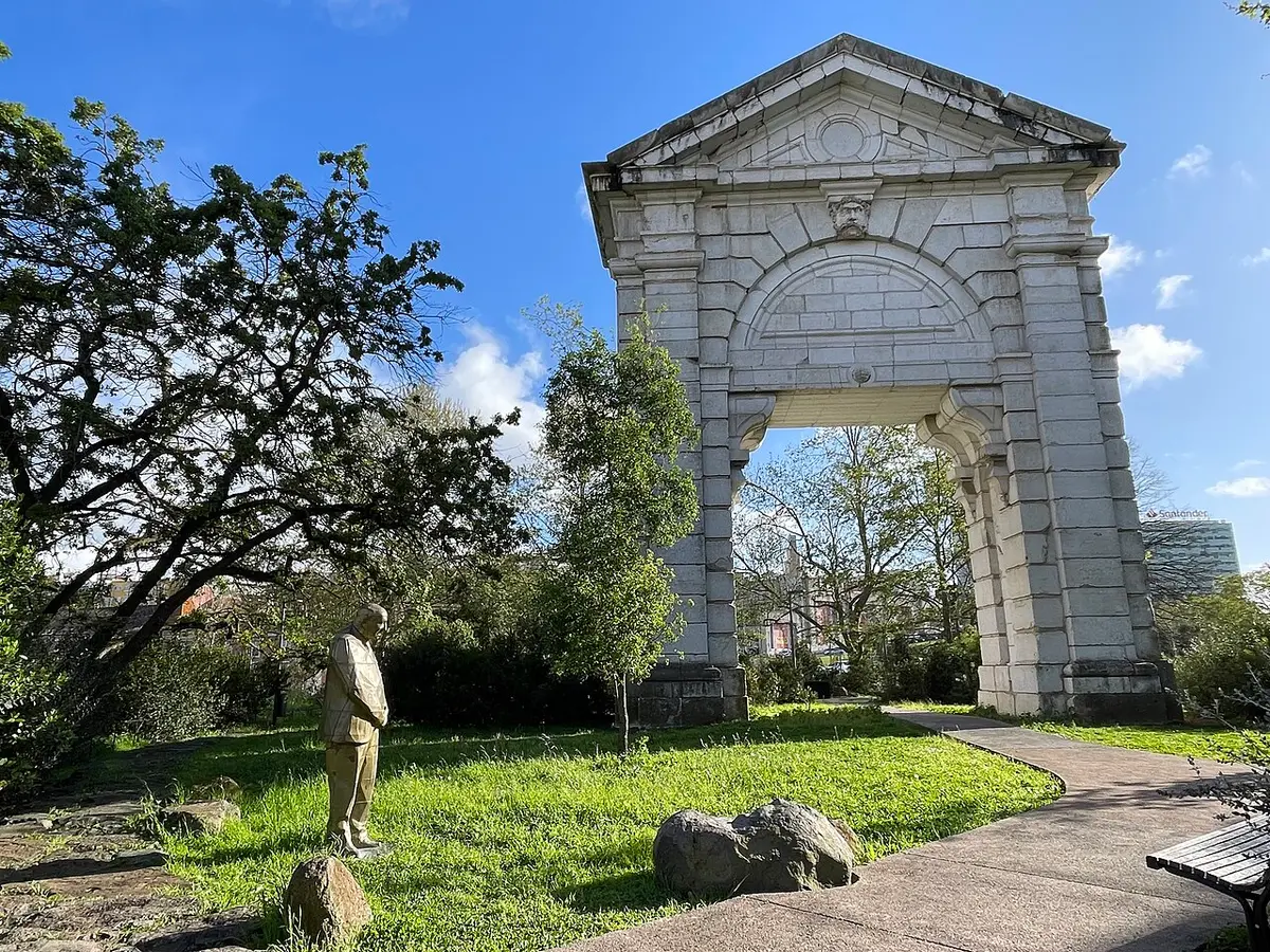 Arco de pedra monumental e estátua num jardim relvado com árvores, sob céu azul