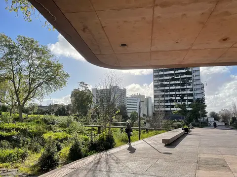 Parque urbano com vegetação, bancos de betão e cobertura de betão, com prédios ao fundo
