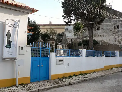 Exterior da Casa-Museu José Pedro, com portão azul, muro branco e amarelo e cartaz identificativo.