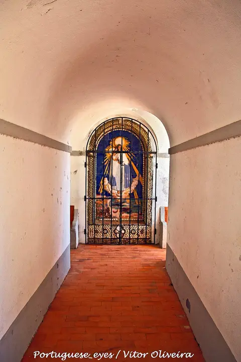 Corredor abobadado branco com chão de tijoleira e painel de azulejos atrás de grade de ferro