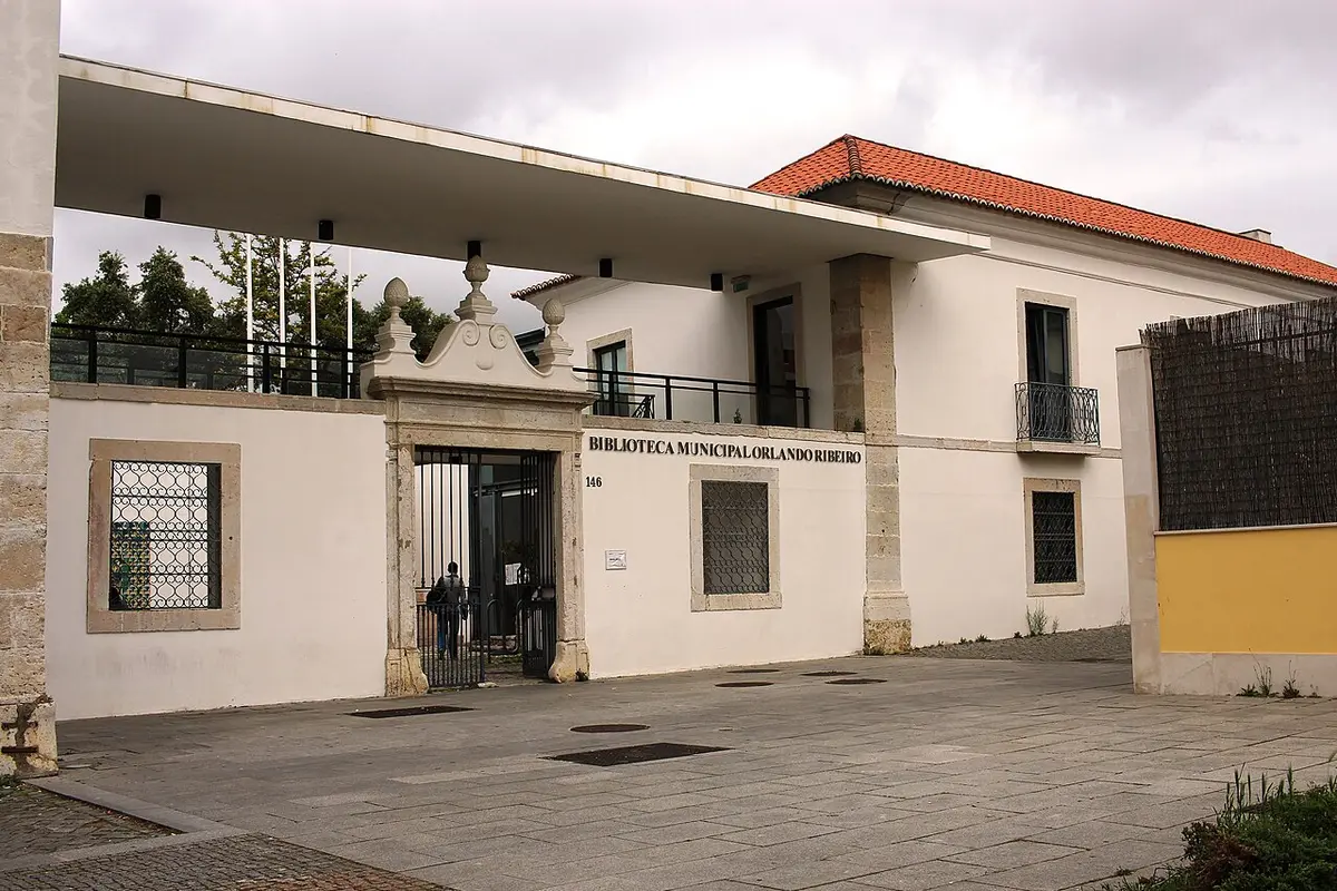 Entrada da Biblioteca Municipal Orlando Ribeiro, em Telheiras, com portão de ferro e letreiro na fachada branca