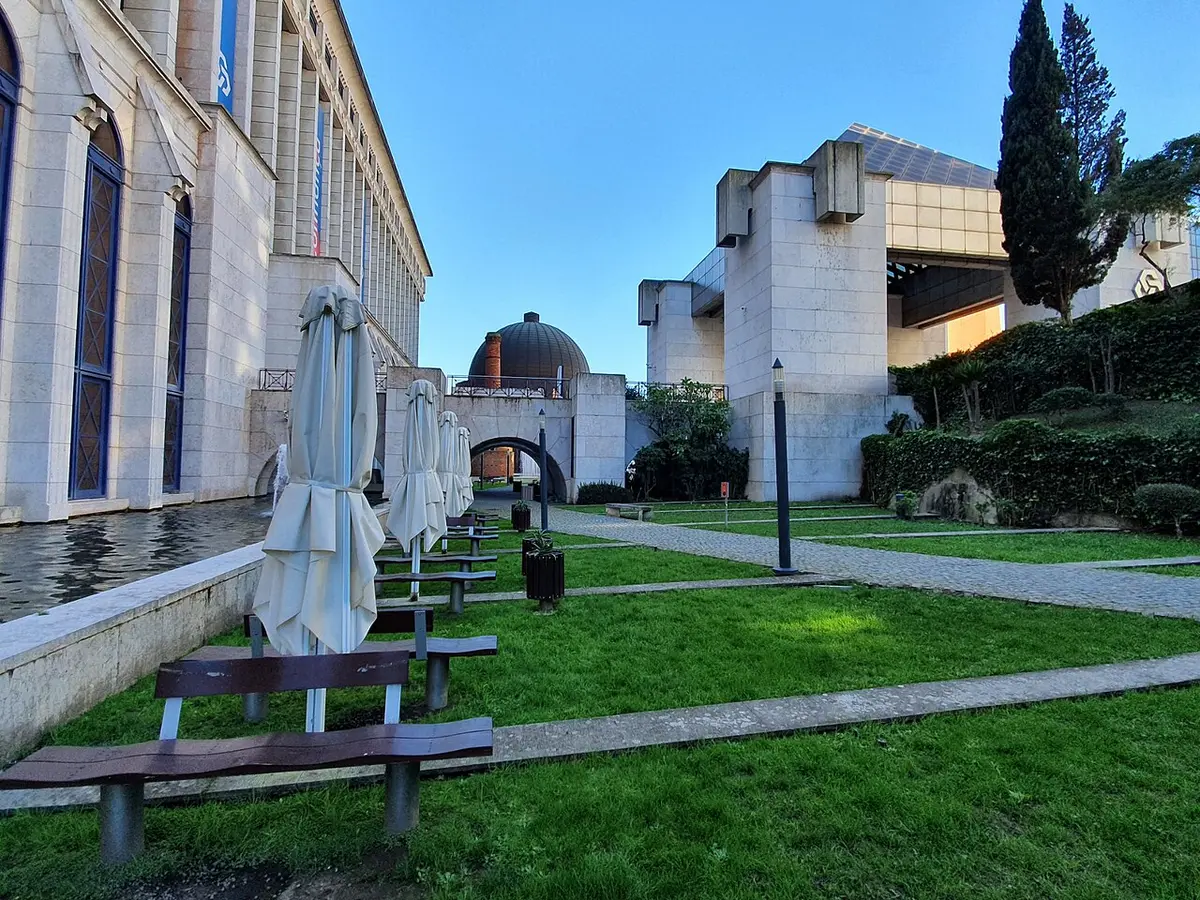 Jardim relvado com bancos e chapéus de sol fechados junto a edifícios em pedra, sob céu azul