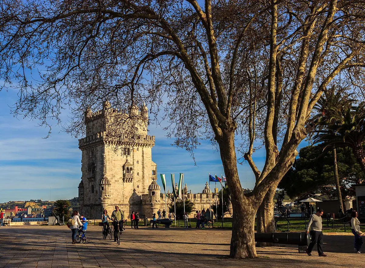 Jardim junto à Torre de Belém, com árvores grandes e pessoas a passear ao fim da tarde