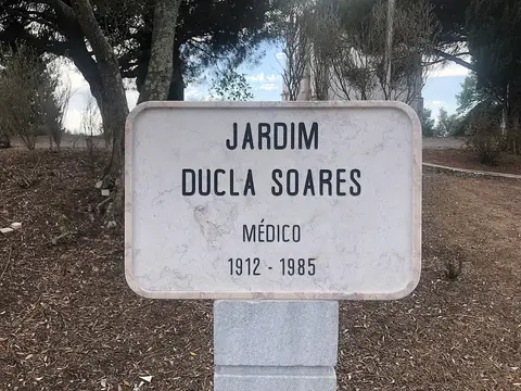 Placa de mármore com a inscrição «Jardim Ducla Soares, médico, 1912-1985» entre árvores.