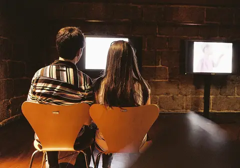 Duas pessoas sentadas de costas a ver ecrãs de vídeo numa sala de pedra do Museu da Imagem, em Braga
