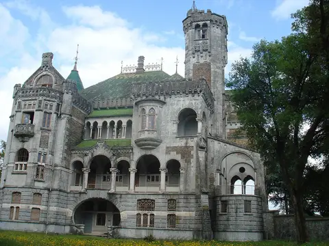 Fachada do Castelo da Dona Chica, com torres, arcadas e telhado verde, rodeado de relva e árvores.