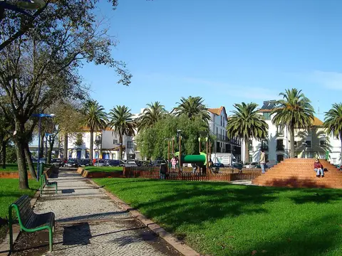 Jardim urbano com relvado, palmeiras, bancos e parque infantil junto a edifícios brancos