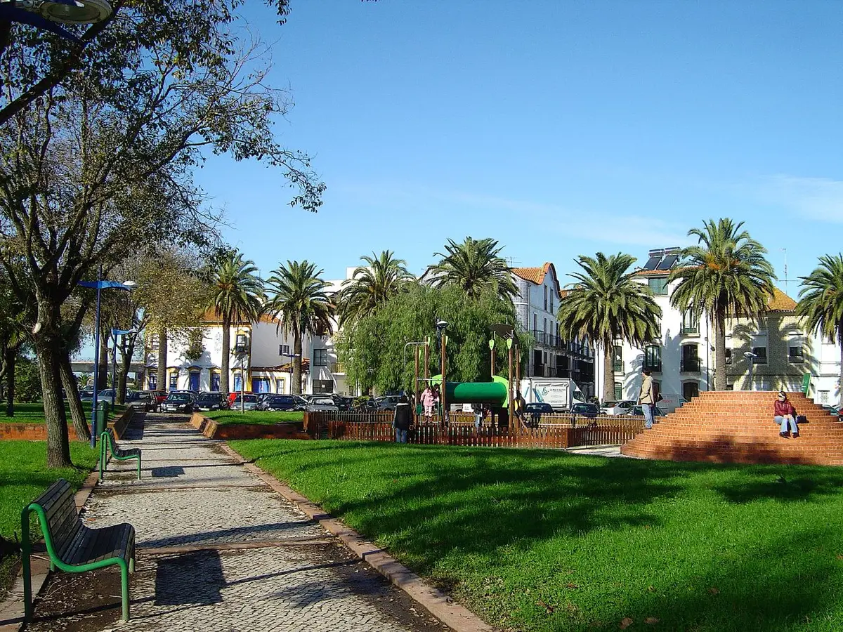 Jardim urbano com relvado, palmeiras, bancos e parque infantil junto a edifícios brancos