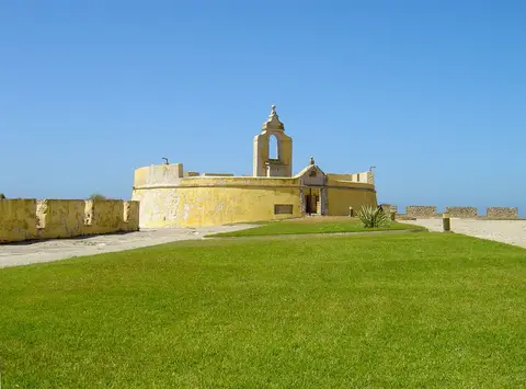 Edifício amarelo com campanário no interior da Fortaleza de Peniche, rodeado de relva sob céu azul