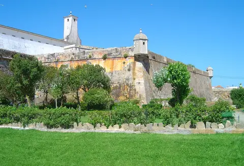 Muralhas e guarita da Fortaleza de Peniche vistas do jardim relvado envolvente, sob céu azul.