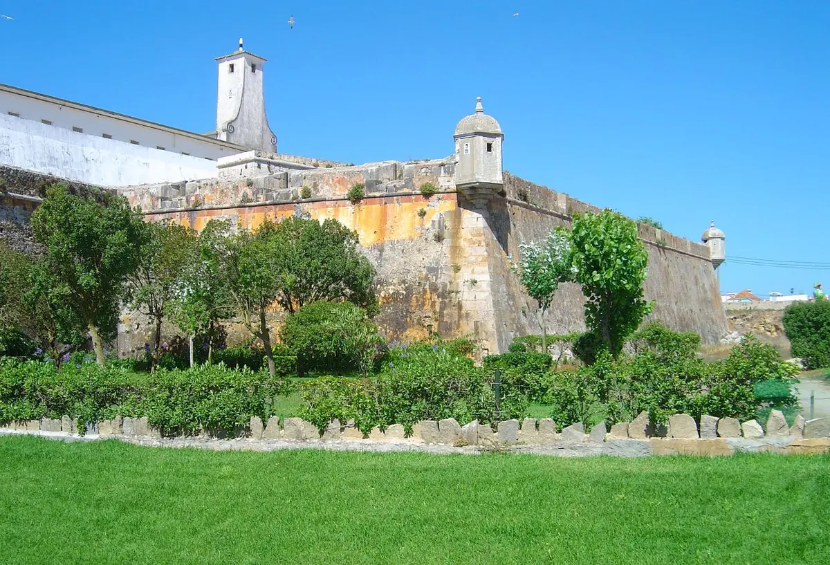 Muralhas e guarita da Fortaleza de Peniche vistas do jardim relvado envolvente, sob céu azul.