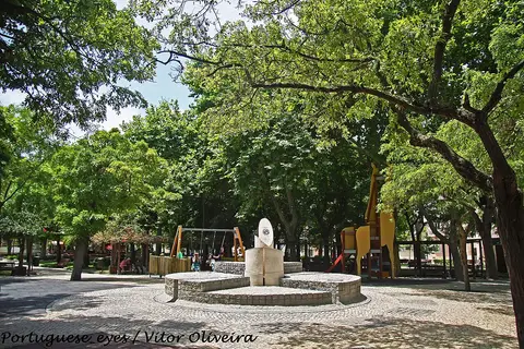 Parque arborizado com escultura em pedra, bancos e parque infantil colorido ao fundo