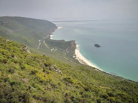 Vista do alto da serra da Arrábida sobre a encosta verde, praias de areia e mar turquesa.