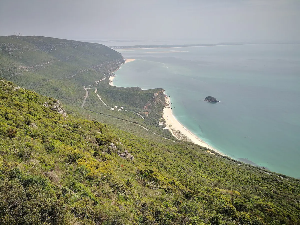 Vista do alto da serra da Arrábida sobre a encosta verde, praias de areia e mar turquesa.
