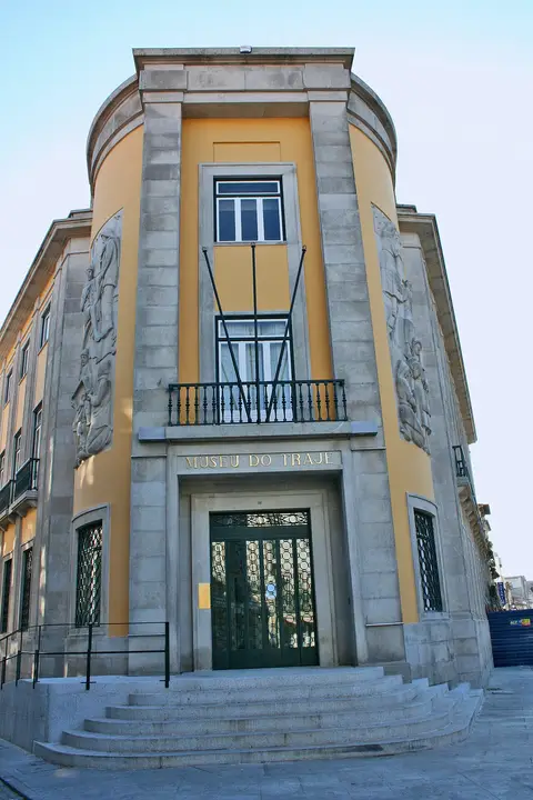 Fachada amarela e em pedra do Museu do Traje, com letreiro sobre a porta e escadaria de acesso.
